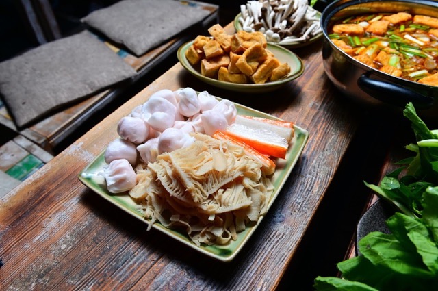 Asiatisches Hotpot-Gericht mit Tofu und Gemüse auf einem Holztisch umgeben von verschiedenen Beilagen.